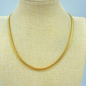 May include: A gold-toned herringbone chain necklace featuring a flat, flexible design with a smooth, polished finish. The necklace consists of tightly woven, interlocking metal links that create a continuous, sleek, and uniform appearance around the neck.