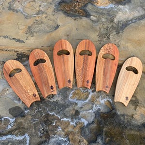May include: Six wooden surfboard-shaped serving trays with a cutout handle. The trays are made of light brown wood with a darker brown wood grain. Each tray has a small star design on the bottom.