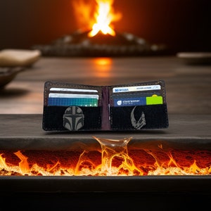 May include: Open brown leather wallet with multiple card slots. The wallet features a black patch with a Mandalorian helmet emblem and a silver emblem. The wallet is open, displaying cards and a driving licence.