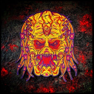 Puede incluir: Parche bordado con una detallada representación de la cara de un Depredador. El diseño utiliza colores vibrantes, incluyendo amarillo, rojo y morado, con intrincadas puntadas. El parche tiene una expresión amenazante y dientes afilados.