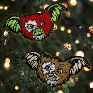 May include: Two embroidered patches of the fictional creature Gizmo from the movie Gremlins. The first patch is green, red, and white with a menacing expression. The second patch is brown, white, and black with a more friendly expression.