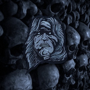 Könnte beinhalten: Ein gestickter Patch in Schwarzweiß, der ein Gesicht mit langen Haaren zeigt. Das Gesicht hat einen ernsten Ausdruck und ist von einem dunklen Hintergrund umgeben.