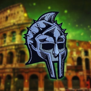 Puede incluir: Parche bordado con un diseño de casco de gladiador. El parche es principalmente negro y gris, con una representación detallada de las características del casco, incluyendo púas y una protección facial. El fondo es una imagen borrosa del Coliseo.