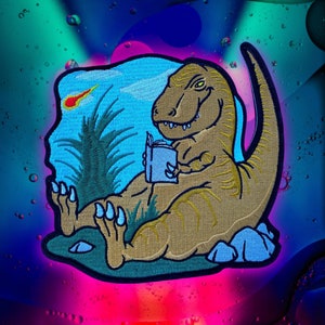 Könnte beinhalten: Ein brauner Dinosaurier mit einem grünen Schwanz sitzt auf einem grünen Grasfleck. Der Dinosaurier hält ein blaues Buch in seinen Händen und hat einen glücklichen Gesichtsausdruck. Ein Komet fliegt im Hintergrund durch den Himmel.