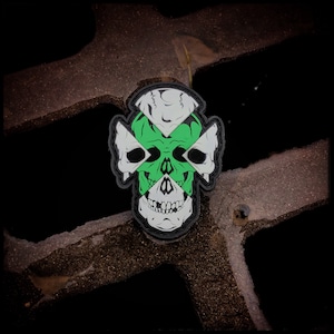 May include: A green, white, and black skull patch with a cross design. The skull is stylized with a cross pattern over the eyes and nose.