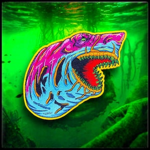 May include: A bright pink and blue embroidered patch of a shark with its mouth open, showing sharp teeth. The shark is set against a green background.