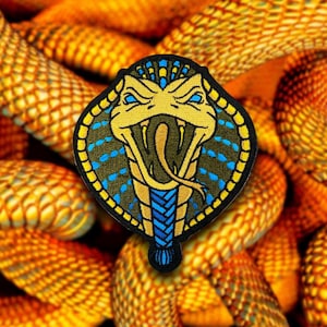 Può includere: Una toppa ricamata con un cobra con la bocca aperta e i denti visibili. Il cobra è giallo e blu, ed è circondato da un cerchio giallo e blu.