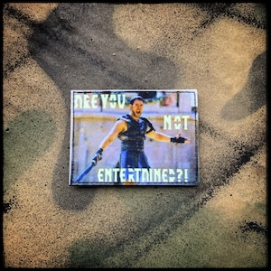 May include: A rectangular patch featuring a scene from a movie with the text "ARE YOU NOT ENTERTAINED?!" in white. The image shows a man in a gladiator outfit with a sword. The patch has a black border and is set against a textured background.