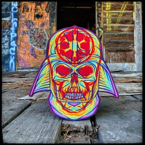 Puede incluir: Un parche bordado colorido con un diseño estilizado del casco de Darth Vader. El parche muestra un vibrante espectro de colores, incluyendo rojo, amarillo, azul y morado, creando un efecto psicodélico. El parche está colocado sobre una superficie de madera envejecida.