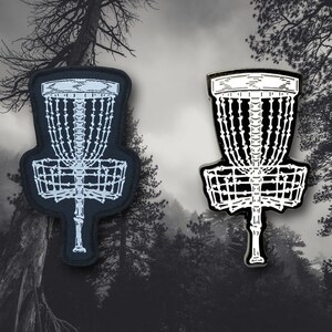May include: Two disc golf basket designs. One is a black patch with white stitching, the other is a black and white enamel pin. Both feature a skeletal design of a disc golf basket, perfect for disc golf enthusiasts.