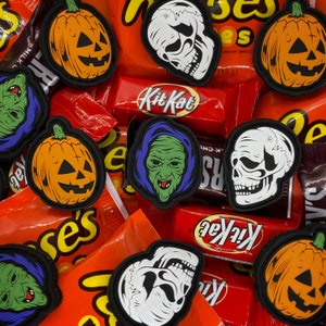 May include: A collection of Halloween-themed iron-on patches featuring pumpkins, skulls, and a monster. The patches are arranged on a background of candy wrappers, including Kit Kat and Reese's.