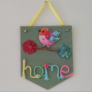 May include: A decorative wooden wall hanging featuring a colourful bird and floral embellishments. The word "home" is spelt out in yarn at the bottom. A yellow ribbon is attached for hanging. The bird is pink, orange, and blue.