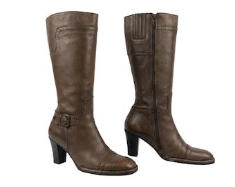 May include: A pair of brown leather knee-high boots. One boot has a buckle closure, and the other has a zipper. Both boots have a block heel and decorative stitching.