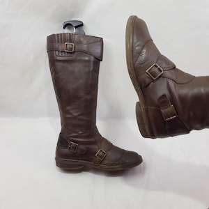 Puede incluir: Un par de botas de cuero marrón de caña alta con hebillas. Las botas tienen punta estrecha y tacón bajo.