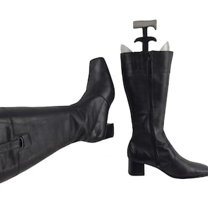 Puede incluir: Botas negras de cuero hasta la rodilla con punta cuadrada y tacón bajo. Una bota está de pie y la otra está tumbada de lado. Hormas insertadas en ambas botas.