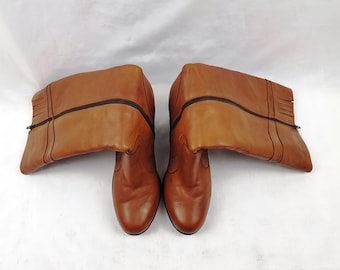 May include: A pair of tan leather boots with a rounded toe and a mid-calf height. Each boot features a dark brown vertical zip running along the outer side, decorative stitching details, and a small fringe accent near the top opening.