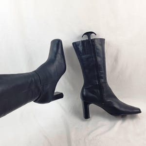 May include: A pair of navy blue leather boots. One boot is held up, showing the shaft and square toe. The other boot is upright, displaying a side zipper and a high heel. The boots are on a white background.