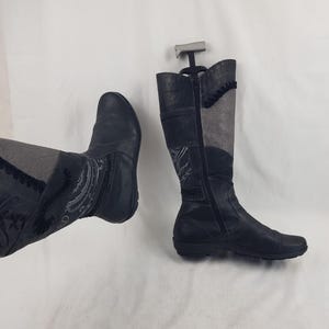 Op de afbeelding: Twee zwarte leren kniehoge laarzen. Eén laars staat rechtop met een rits, de andere staat schuin met een veterdetail. Beide laarzen hebben grijze stoffen panelen en decoratieve stiksels. De laarzen staan op een witte achtergrond.