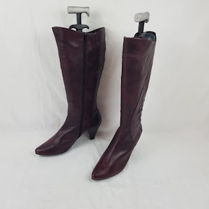 Peut inclure: Une paire de bottes en cuir bordeaux jusqu'aux genoux. Les bottes ont un bout pointu, une fermeture éclair latérale et un petit talon. Les bottes sont présentées sur des embauchoirs sur fond blanc.