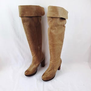 Puede incluir: Un par de botas altas de ante color canela con un puño doblado. Las botas tienen una punta redondeada, una suela de plataforma y un tacón apilado. Una cremallera recorre el interior de cada bota. Los tacones miden aproximadamente 7.6 cm de alto.