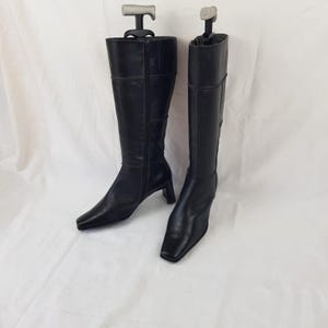 May include: A pair of black knee-high boots featuring a square toe, a low block heel, and a full-length side zipper. The boots are supported by grey plastic boot trees inserted into the shafts to maintain their upright structure.