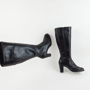 May include: A pair of black leather knee-high boots. One boot is lying on its side, while the other stands upright. Both boots have a heel and a zip closure. The boots are set against a white background.