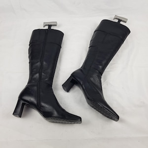 May include: A pair of black knee-high boots featuring a square toe, a block heel, and a full-length side zipper. The boots are constructed with visible stitched paneling along the shaft and include grey plastic boot trees inserted at the top.