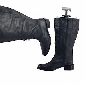 Puede incluir: Botas altas de cuero negro con diseño de cordones y cremallera. Un horma de bota está insertada en una bota. Las botas están sobre un fondo blanco. Las botas son de estilo clásico.