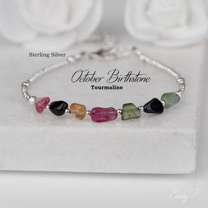 May include: A sterling silver bracelet featuring a series of small, polished, multi-coloured tourmaline gemstone chips in shades of pink, green, black, and amber, interspersed with tiny silver beads. The text reads Sterling Silver October Birthstone Tourmaline.