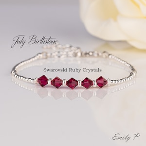 May include: A silver bracelet featuring a line of red Swarovski ruby crystals. The bracelet is displayed against a white background with the text "July Birthstone" and "Swarovski Ruby Crystals".