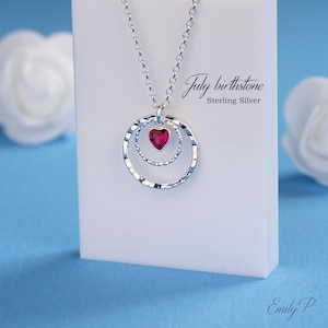 May include: A silver necklace with a heart-shaped July birthstone pendant. The pendant is set within two textured silver circles. The necklace is presented on a white surface against a blue background. The text "July birthstone Sterling Silver" is visible.