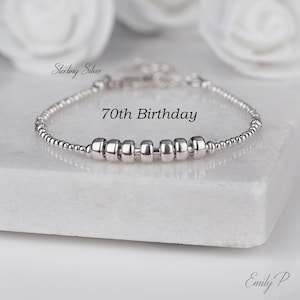 May include: A sterling silver bracelet with the text "70th Birthday". The bracelet features small, round beads and larger, cylindrical beads. The bracelet is displayed on a white surface with white flowers in the background.