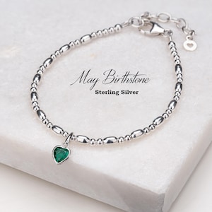 May include: A sterling silver bracelet featuring a green heart charm. The bracelet is made of small, round beads and has a clasp fastening. The charm is a heart-shaped pendant with a green gemstone.