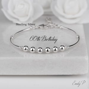 May include: A silver bracelet featuring six spherical beads. The text "60th Birthday" is displayed. The bracelet is presented on a white surface, with white flowers in the background. The words "Sterling Silver" are also visible.