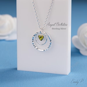 May include: A sterling silver necklace featuring two interlocking hammered circular pendants and a small heart-shaped green peridot gemstone. The display card includes the text August Birthstone Sterling Silver and the signature Emily P.