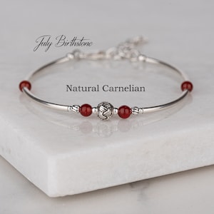 May include: A silver bracelet with a red carnelian gemstone and a silver charm. The bracelet is on a white marble surface. "July Birthstone" and "Natural Carnelian" are written in the image.