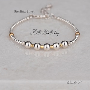 May include: A sterling silver bracelet with the text "50th Birthday." The bracelet features silver beads and gold accents. The bracelet is delicate and elegant, suitable for a special occasion.