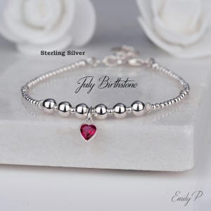 May include: A silver bracelet with a July birthstone heart charm. The bracelet has small silver beads and larger, polished silver beads. The heart-shaped charm is a deep red colour. The words "Sterling Silver" and "July Birthstone" are visible.