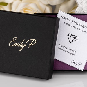 K&ouml;nnte beinhalten: Eine schwarze Geschenkbox mit goldener Beschriftung "Emily P" auf der Vorderseite. Die Box ist ge&ouml;ffnet und im Inneren befindet sich eine wei&szlig;e Karte mit dem Text "HAPPY 60TH BIRTHDAY! 6 Beads For 6 Decades" und einem Diamanten-Symbol. Die Karte tr&auml;gt au&szlig;erdem die Aufschrift "STERLING SILVER Handmade In England" und "Emily P".
