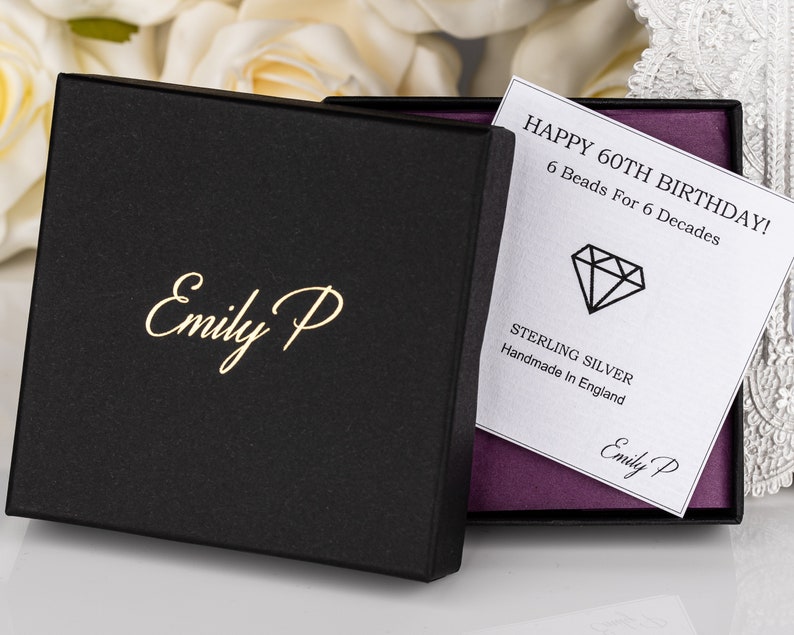 K&ouml;nnte beinhalten: Eine schwarze Geschenkbox mit goldener Beschriftung "Emily P" auf der Vorderseite. Die Box ist ge&ouml;ffnet und im Inneren befindet sich eine wei&szlig;e Karte mit dem Text "HAPPY 60TH BIRTHDAY! 6 Beads For 6 Decades" und einem Diamanten-Symbol. Die Karte tr&auml;gt au&szlig;erdem die Aufschrift "STERLING SILVER Handmade In England" und "Emily P".