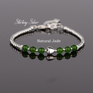 May include: A sterling silver bracelet with a heart charm and green jade beads. The bracelet is on a reflective surface.