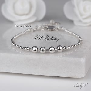 May include: A silver bracelet with the inscription "40th Birthday". The bracelet is made of small, round beads and larger, polished silver beads. The bracelet is presented on a white surface.