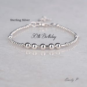 May include: A silver bracelet featuring small beads and five larger, spherical beads. The text "50th Birthday" is displayed on the bracelet. The words "Sterling Silver" and "Emily P" are also visible.