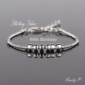 May include: A sterling silver bracelet featuring a series of small, round beads along the chain and six larger, cylindrical beads centred on a thin bar. The text "Sterling Silver" and "60th Birthday" are visible above the bracelet.