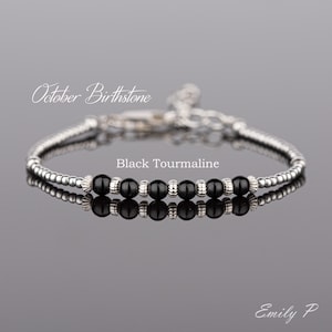 May include: October birthstone bracelet featuring black tourmaline beads and silver beads. The bracelet is displayed on a reflective surface. The text "Black Tourmaline" is written below the bracelet.