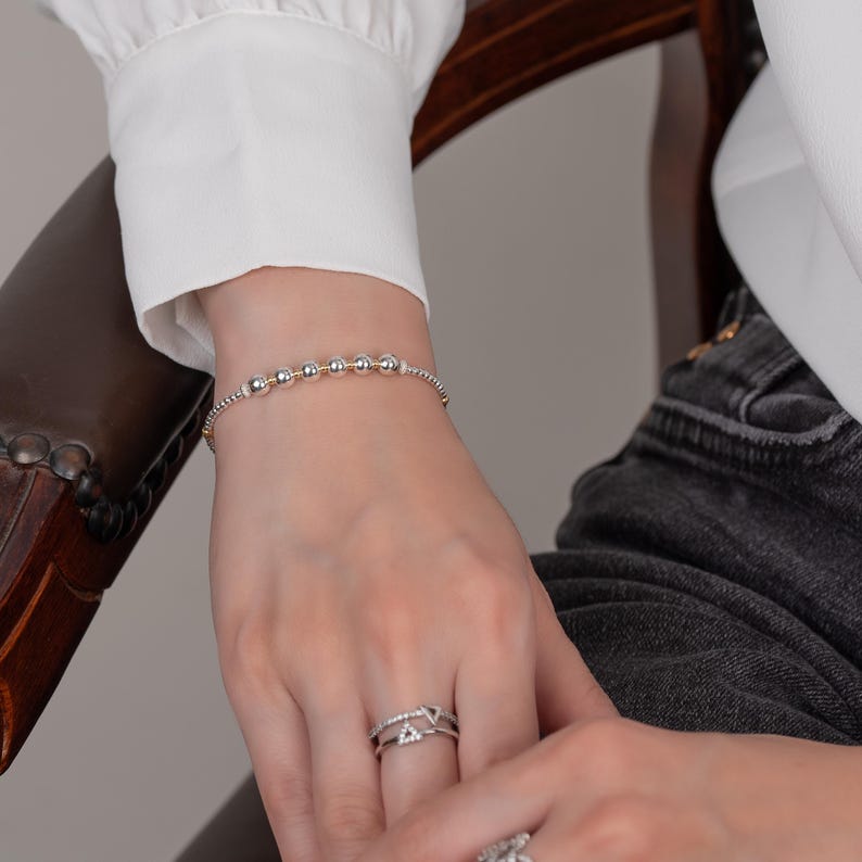 K&ouml;nnte beinhalten: Ein silbernes und goldenes Armband mit Perlen und einer zarten Kette. Das Armband befindet sich an einem Handgelenk.