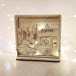 Könnte beinhalten: Eine quadratische Holzlichtbox mit einer mehrschichtigen, lasergeschnittenen Stadtszene, einem Oldtimer, einem Motelschild und einem Café mit der Aufschrift COFFEE, OPEN und GOOD COFFEE GOOD DAYS. Die Szene wird von innen warm beleuchtet.