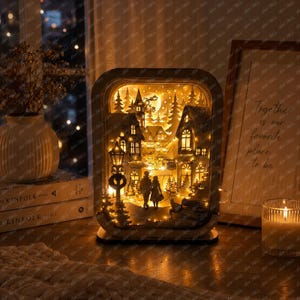 May include: A rectangular wooden light box featuring a layered, laser-cut winter village scene. The illuminated display shows silhouettes of two figures walking through a town with houses, trees, a street lamp, and a bench under a starry sky.