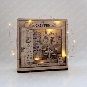 May include: A square wooden light box featuring an engraved coffee shop interior scene with a door, table, chairs, and menu. The text reads COFFEE, OPEN, MENU, WELCOME, and BUT FIRST COFFEE. Warm fairy lights are wrapped around the frame.