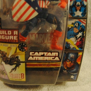 May include: Captain America action figure in packaging. The figure is wearing a blue and red costume with a shield. The packaging includes the text "Captain America", "Build a Figure Collection", and "Marvel".